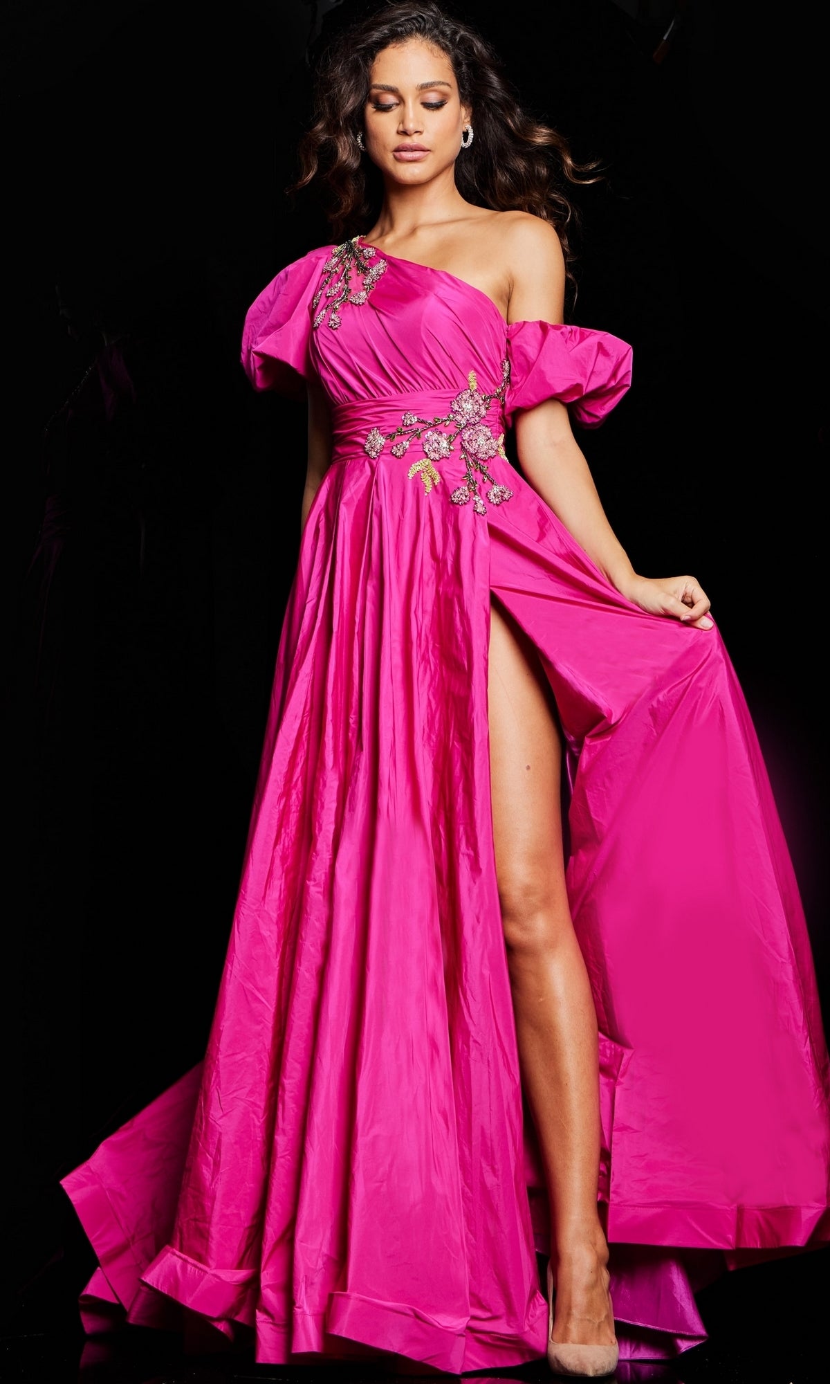 A woman models Jovani’s Long Prom Dress 36872, a bright pink off-the-shoulder gown with floral details, puffed sleeves, and a high slit, posed against a black background.